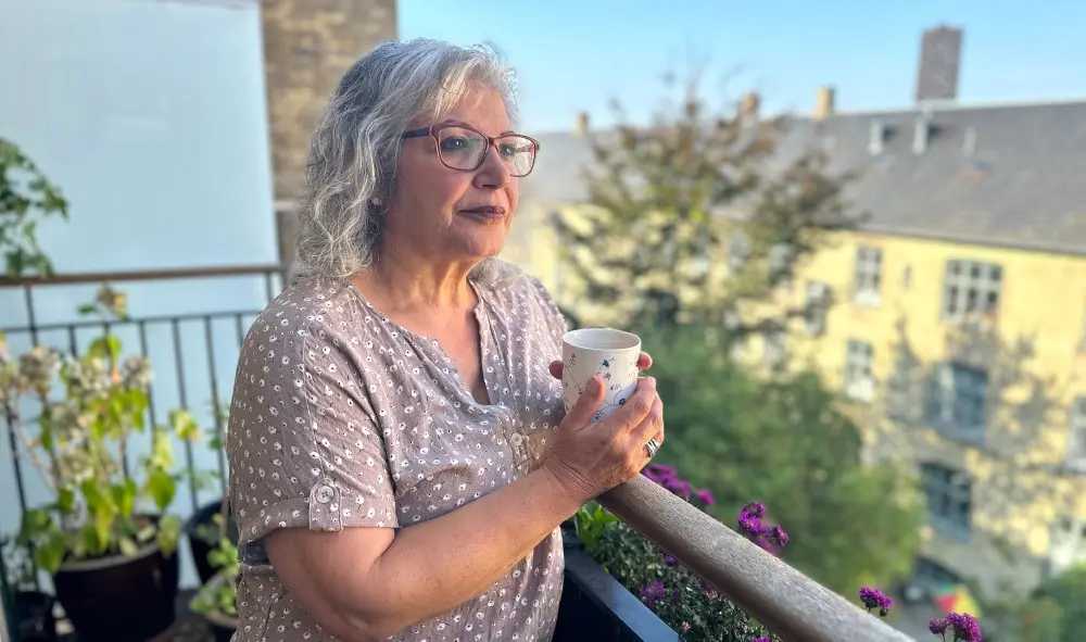 Gül der står på en altan med en kop kaffe i hænderne og kigger ud over gården